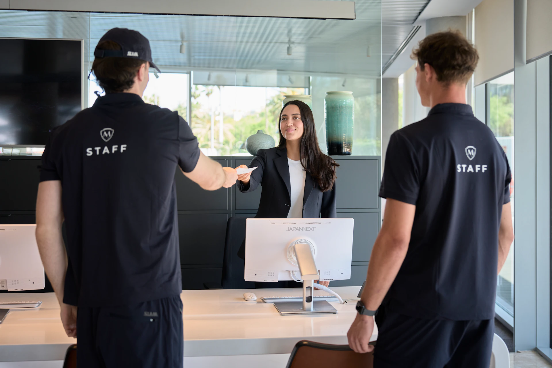 Receptionist hands a card to two staff members in navy uniforms at a front desk.