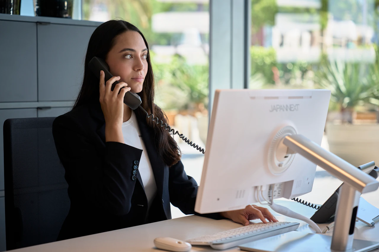 Woman in a blazer talking on a phone at an office desk with a monitor.