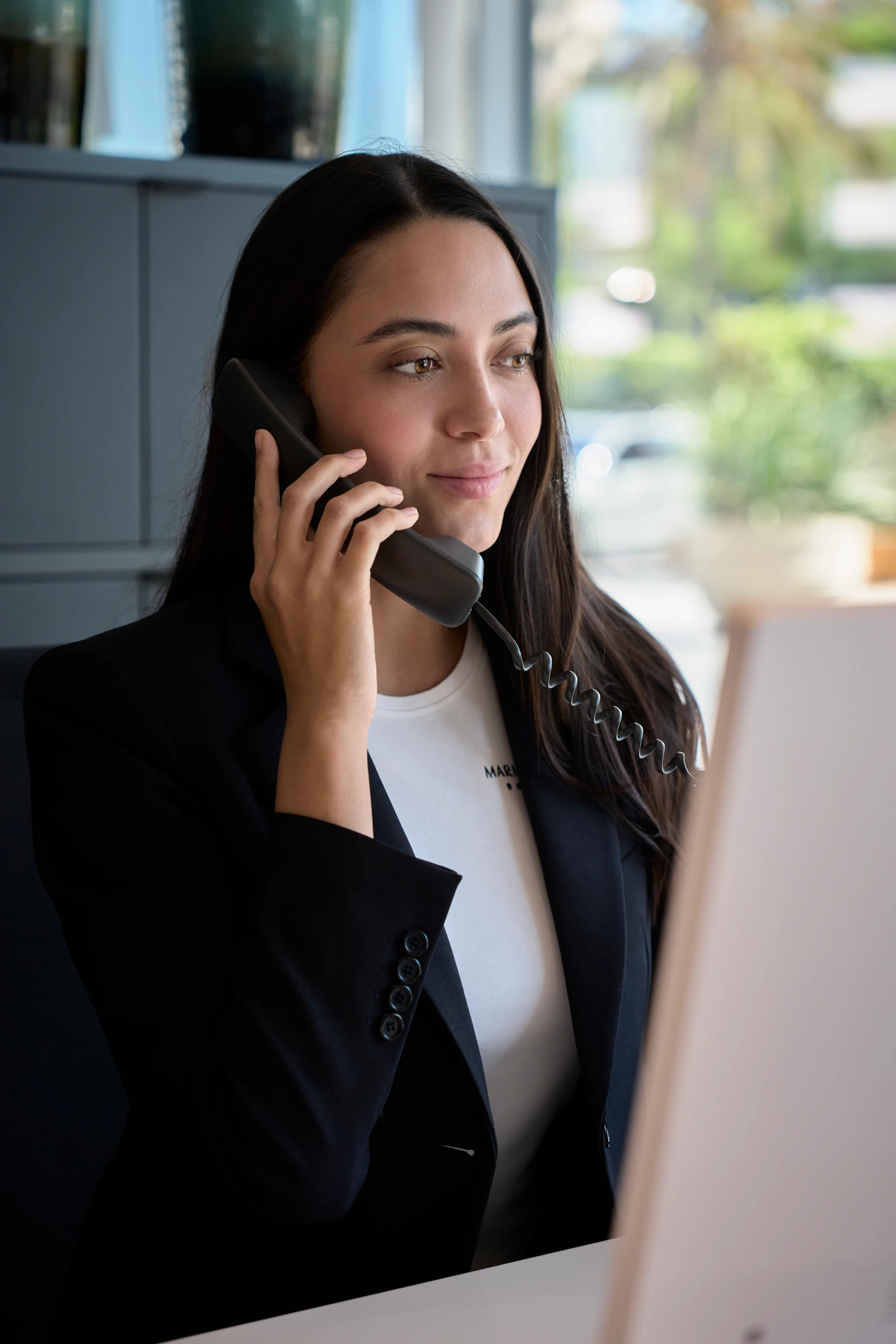 Mujer con blazer negro hablando por un teléfono con cable en un escritorio.