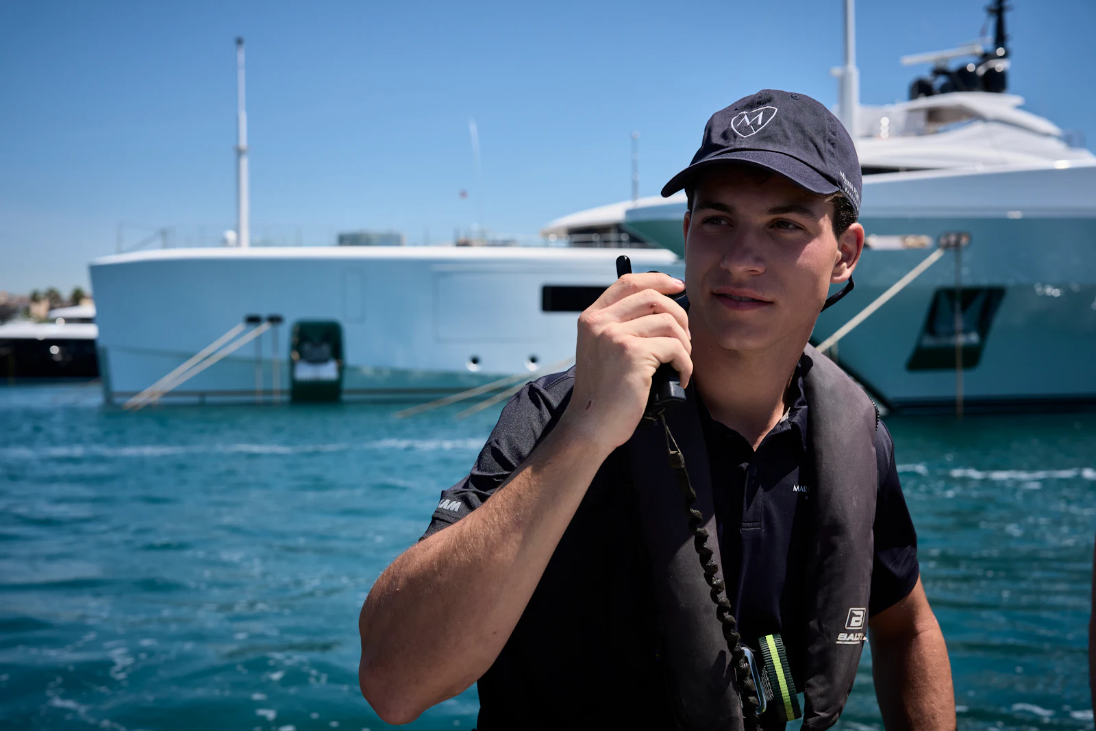 Trabajador de la marina con gorra y chaleco salvavidas sujetando una radio frente a un superyate.