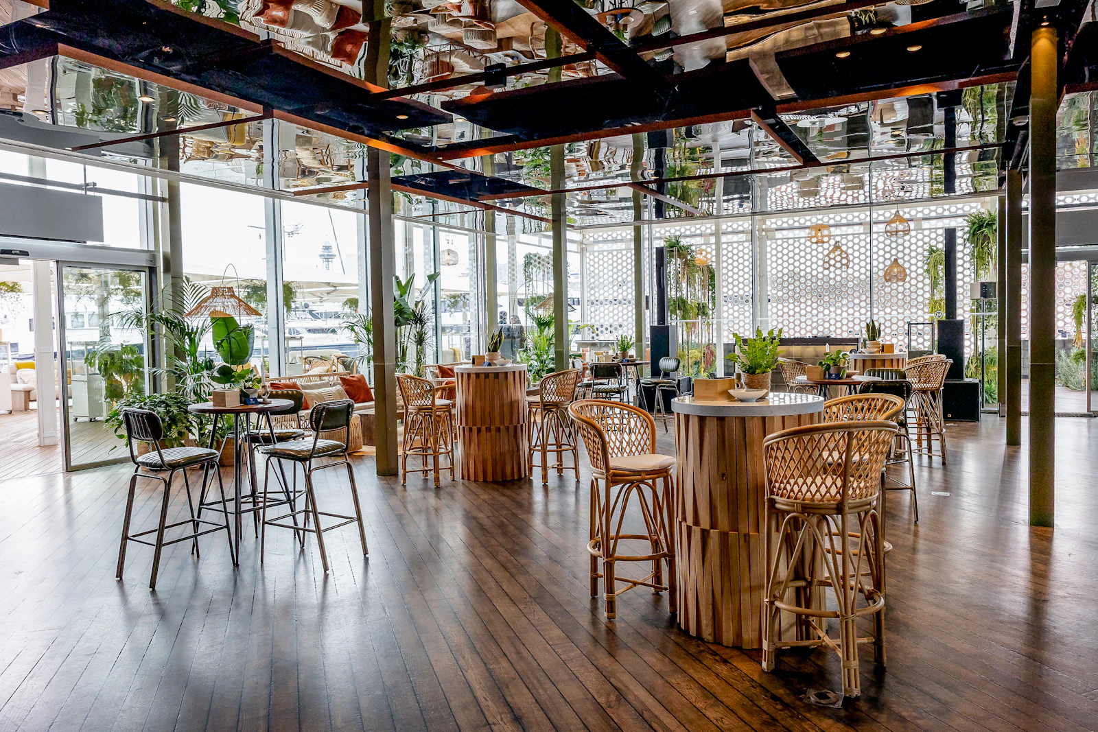 Bright restaurant interior with rattan bar stools, wooden tables and plants by large windows.