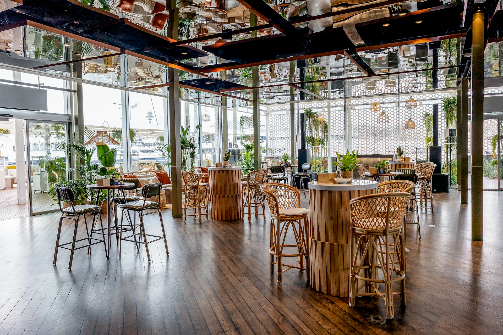 Bright restaurant interior with rattan bar stools, wooden tables and plants by large windows.