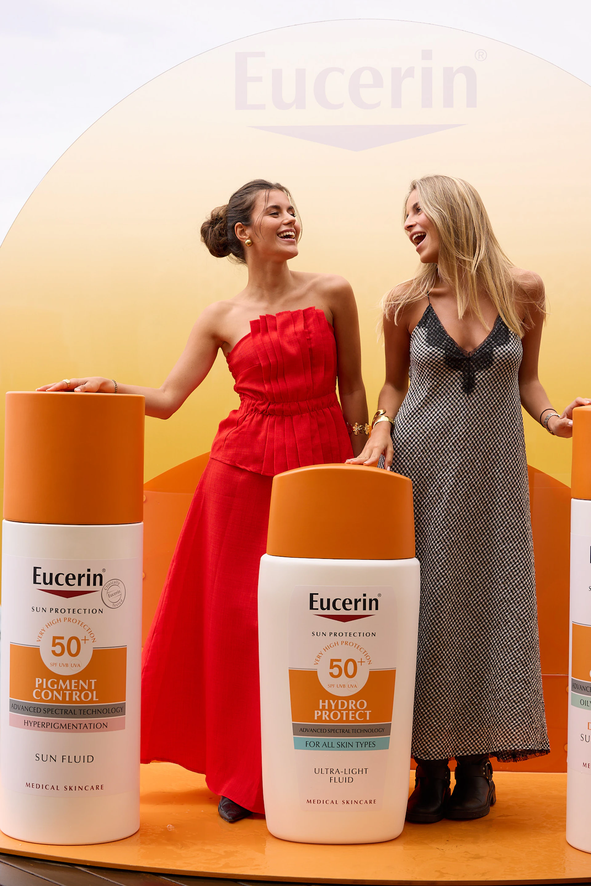 Two women laughing beside giant Eucerin sunscreen bottles at a branded event display.