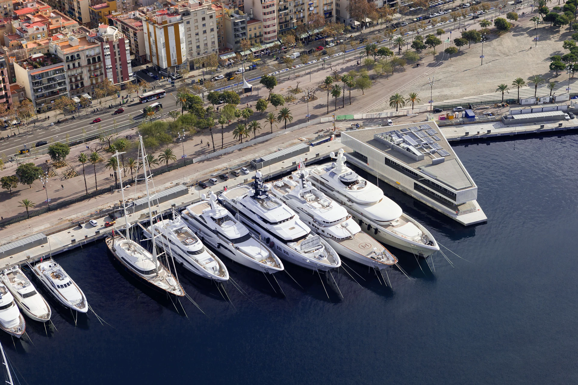 Multiple superyachts moored at Marina Port Vell with Barcelona city buildings and beachfront in the background.