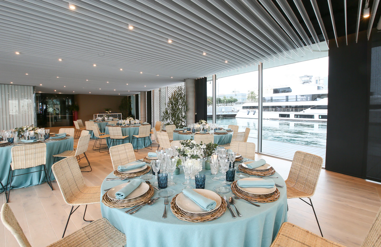 Elegante espacio para eventos con mesas redondas vestidas con mantelería azul claro, sillas de mimbre y servicios de mesa con vistas a un puerto deportivo con yates amarrados.