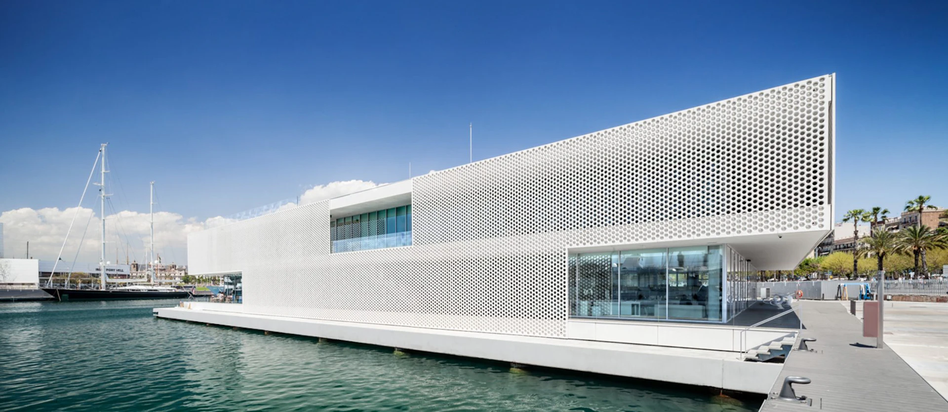 Edificio blanco moderno con fachada de metal perforado en el paseo marítimo de Barcelona junto a un yate amarrado.