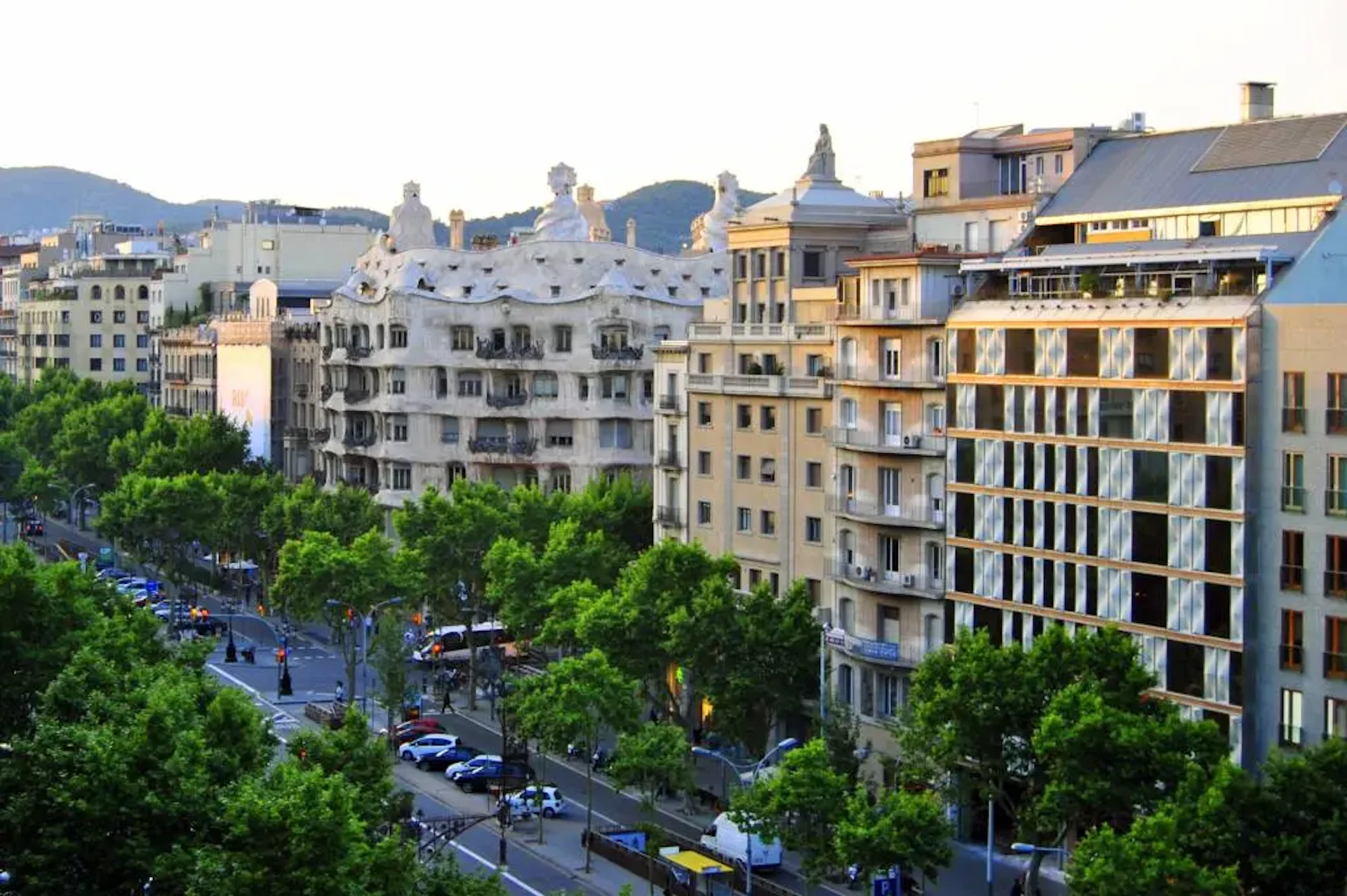 Paisaje urbano de Barcelona con una avenida arbolada, edificios modernistas y montañas al fondo.