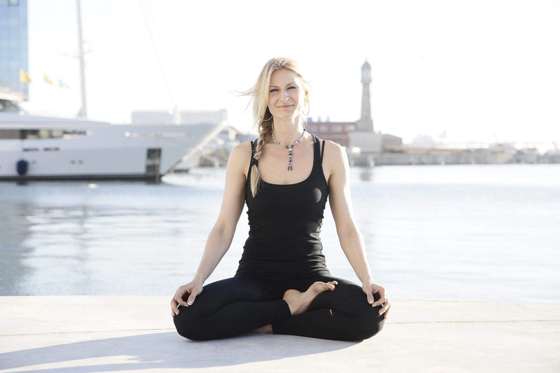 Mujer sentada en posición de meditación con las piernas cruzadas en un muelle junto al agua con superyates amarrados y el perfil de Barcelona al fondo.
