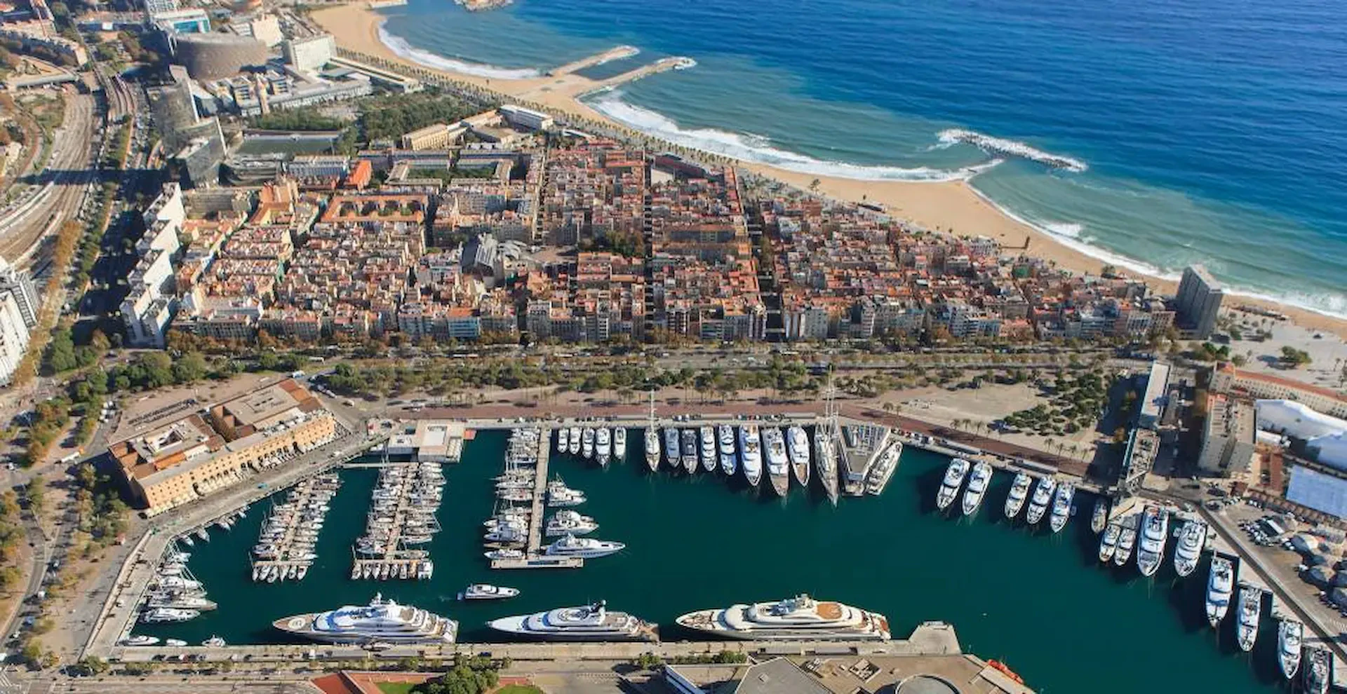 Vista aérea de Marina Port Vell con numerosos superyates amarrados, la playa de Barcelona y el mar Mediterráneo al fondo.
