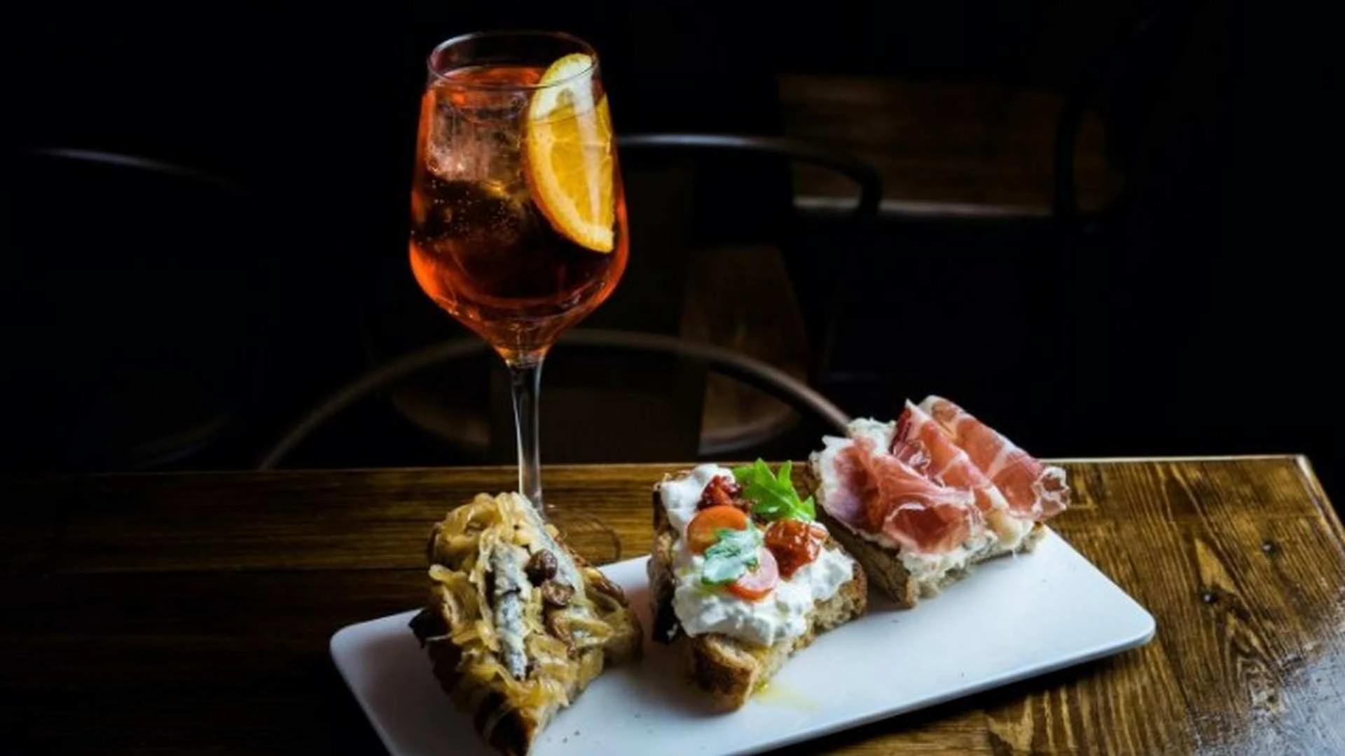 Cóctel con rodaja de naranja junto a un plato de tostas gourmet como aperitivo sobre una mesa de madera.