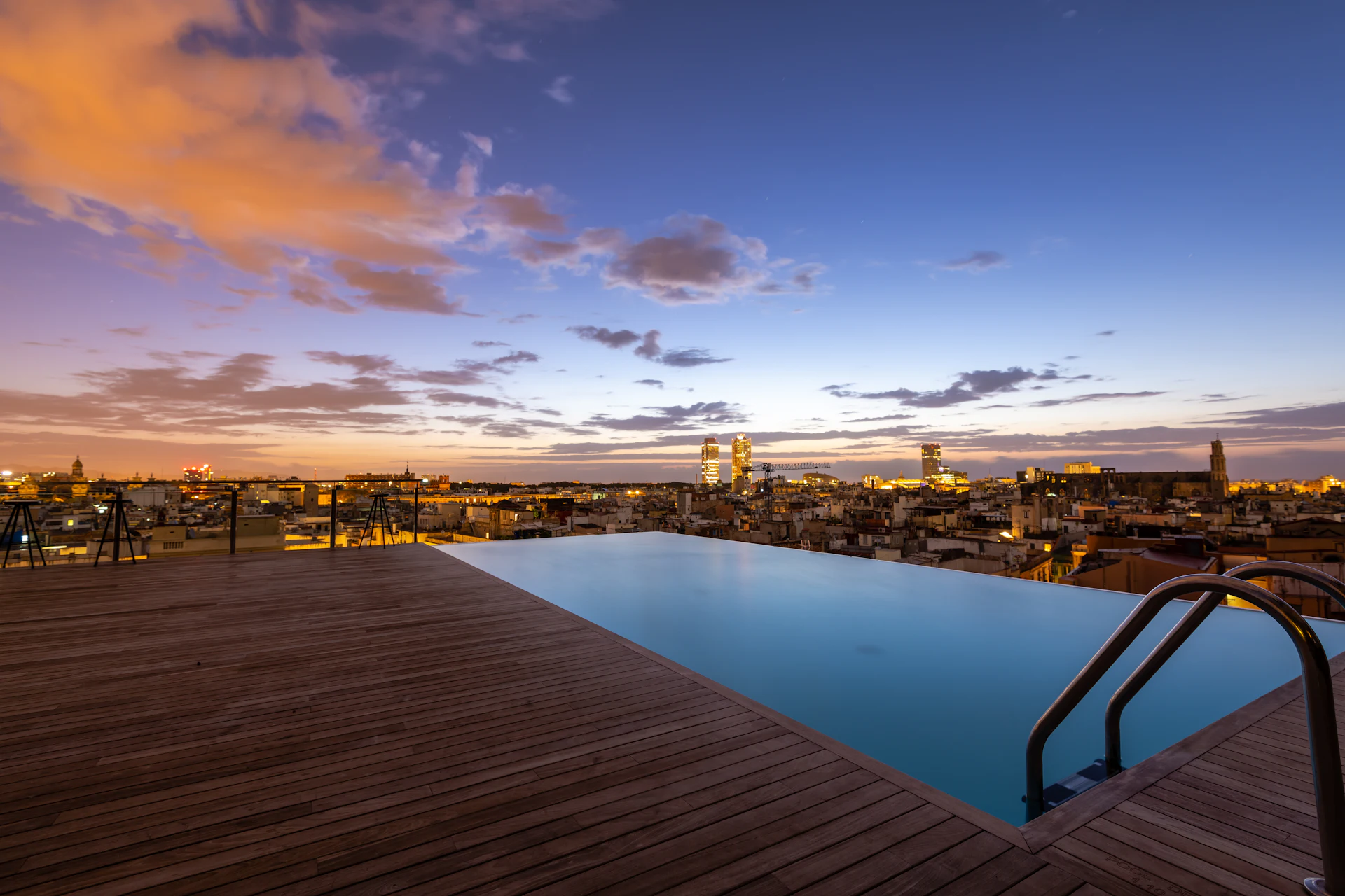 Piscina infinita en la azotea con vistas al paisaje urbano de Barcelona al anochecer con edificios iluminados.