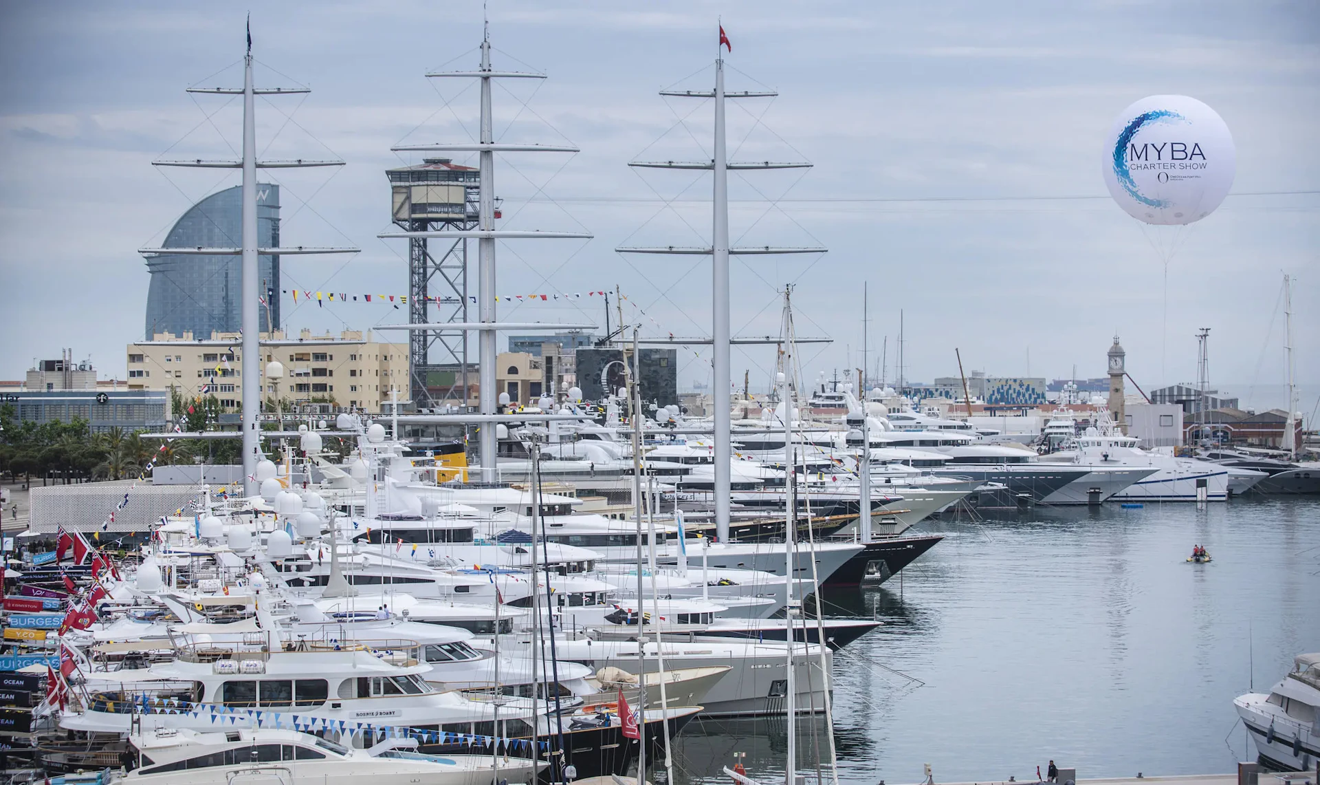 Multiple white superyachts moored at Marina Port Vell in Barcelona with tall masts and city skyline in background.