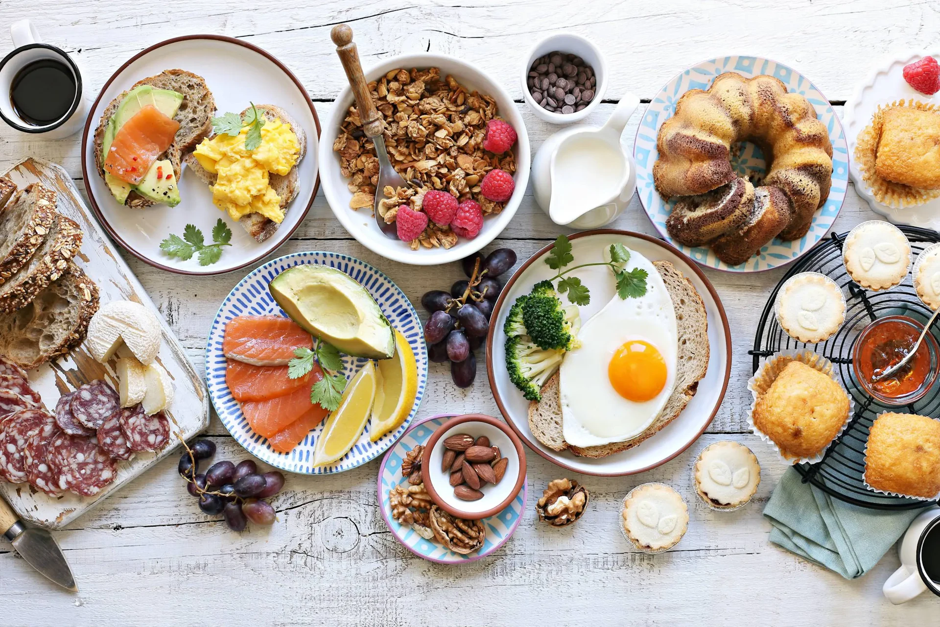 Overhead view of a breakfast spread with toast, eggs, granola, pastries, cured meats, and fresh fruit on white wooden table.