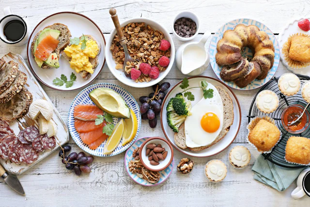 Vista cenital de un desayuno con tostadas, huevos, granola, bollería, embutidos y fruta fresca sobre una mesa de madera blanca.
