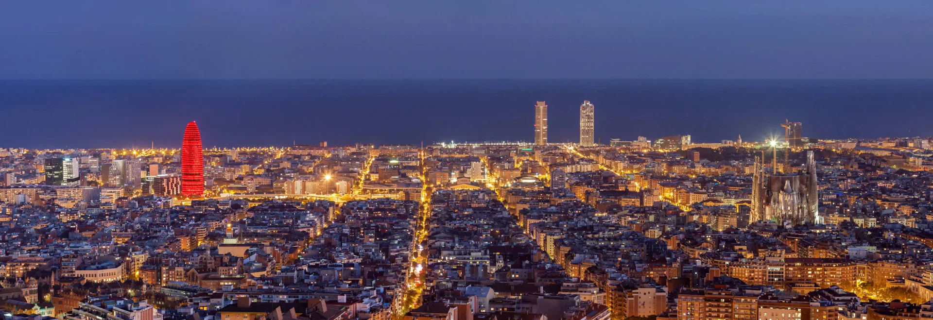 Paisaje urbano de Barcelona al anochecer con la Torre Agbar, la Sagrada Familia y el horizonte costero iluminados.