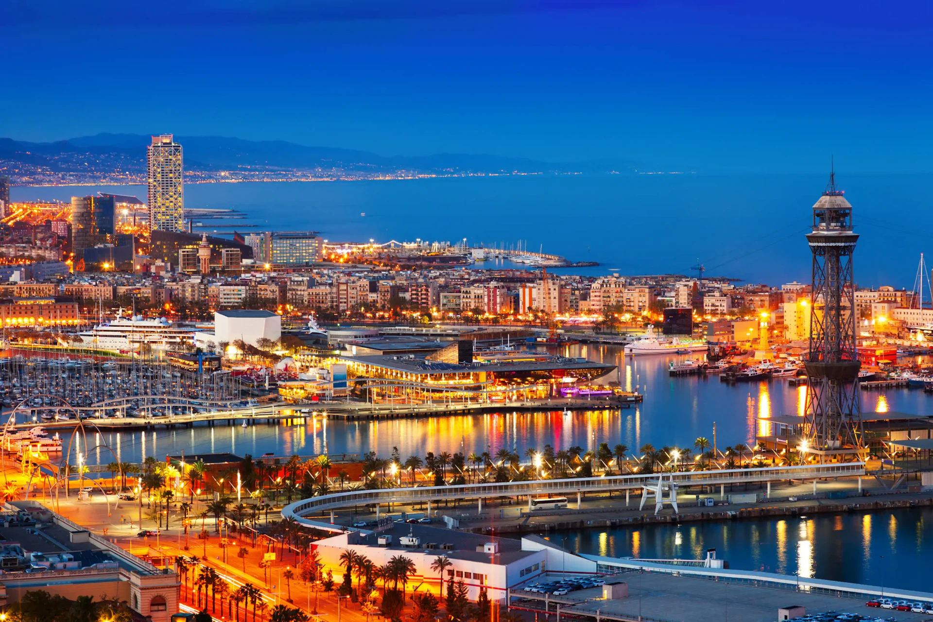 Barcelona waterfront at twilight with illuminated marina, buildings, and cable car tower.