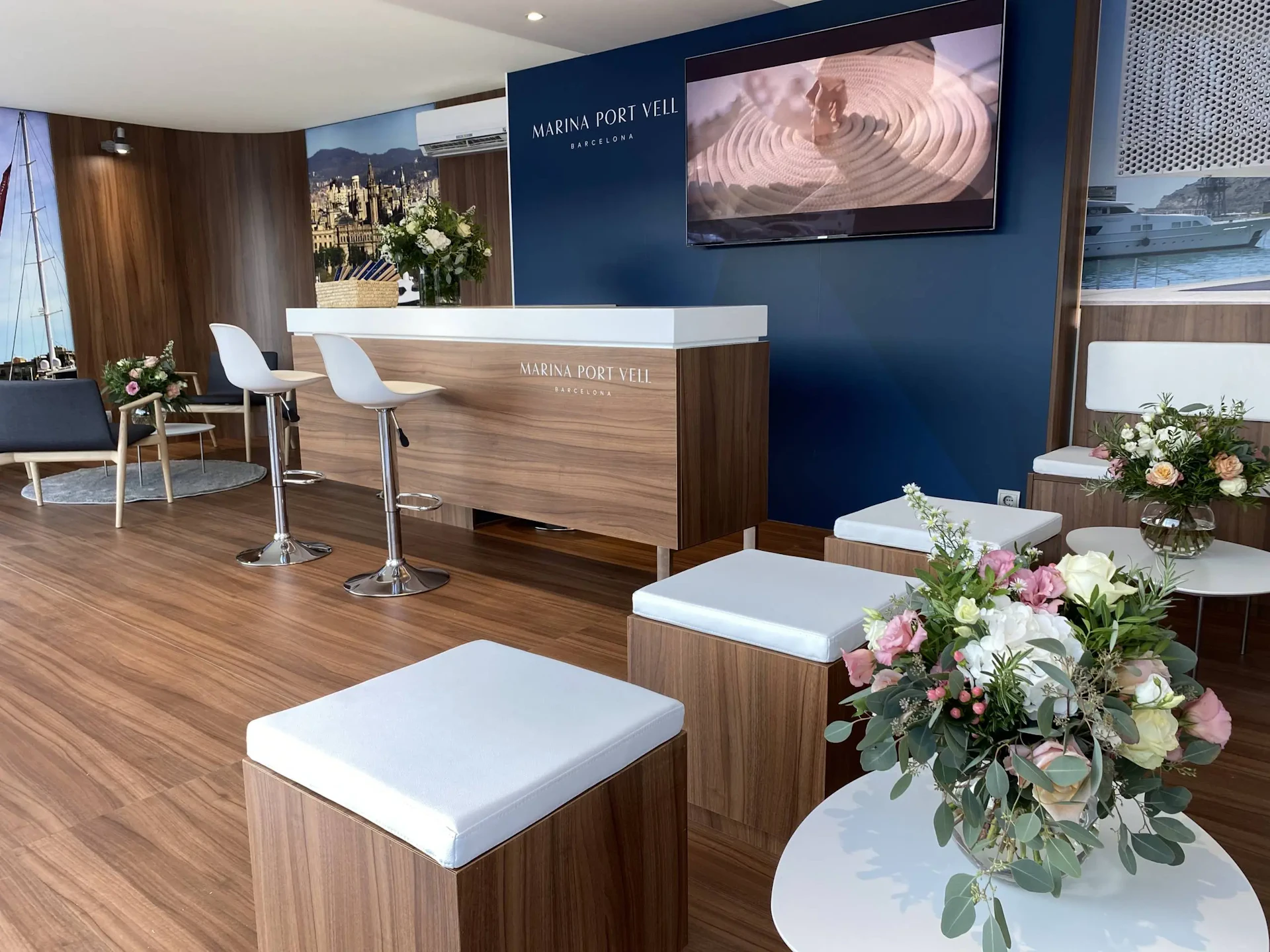 Modern reception area with wooden furniture, white seating, floral arrangements, and Marina Port Vell branding on blue wall.