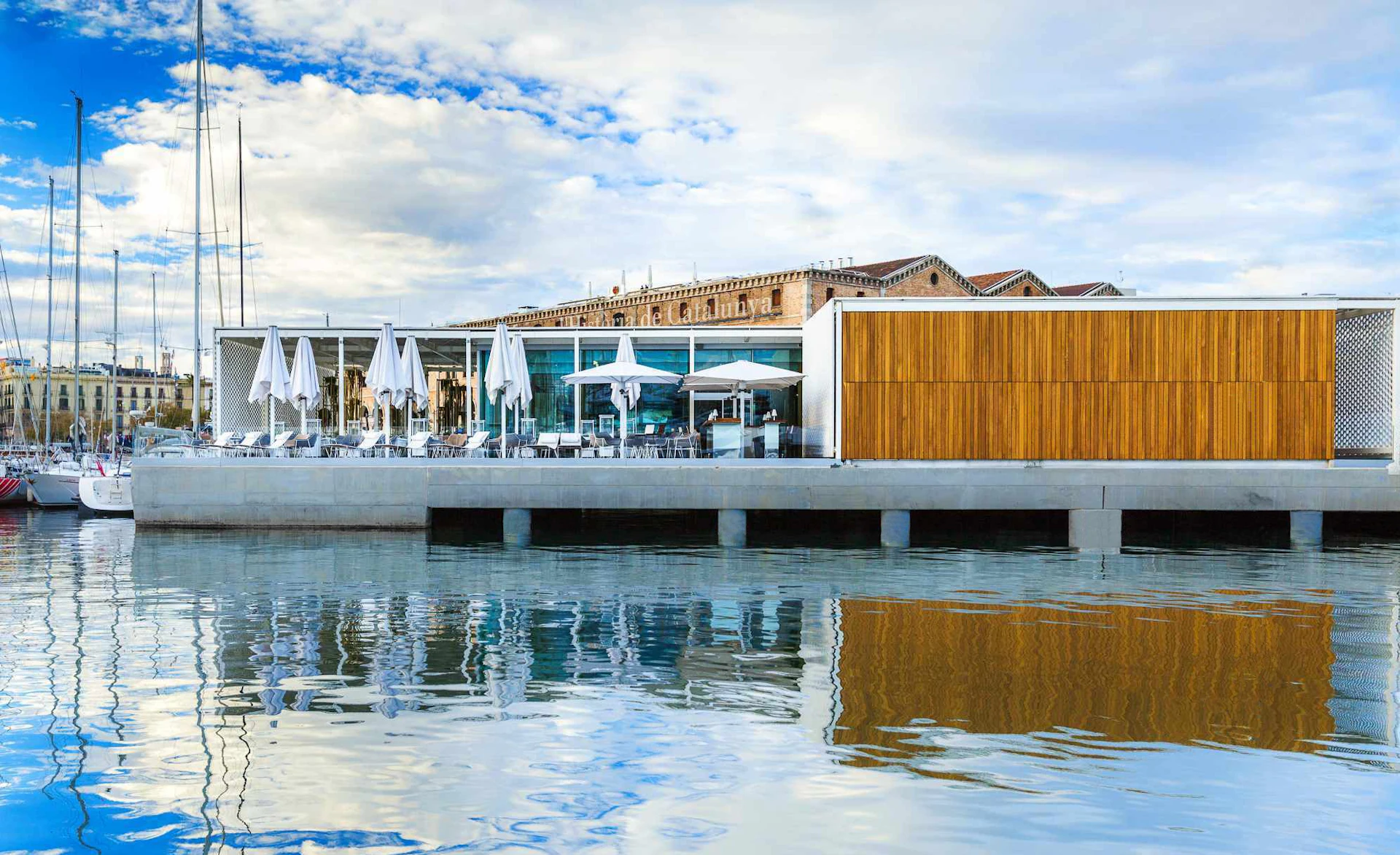 Restaurante y bar modernos junto al agua con sombrillas blancas con vistas a los veleros amarrados en el puerto de Barcelona.