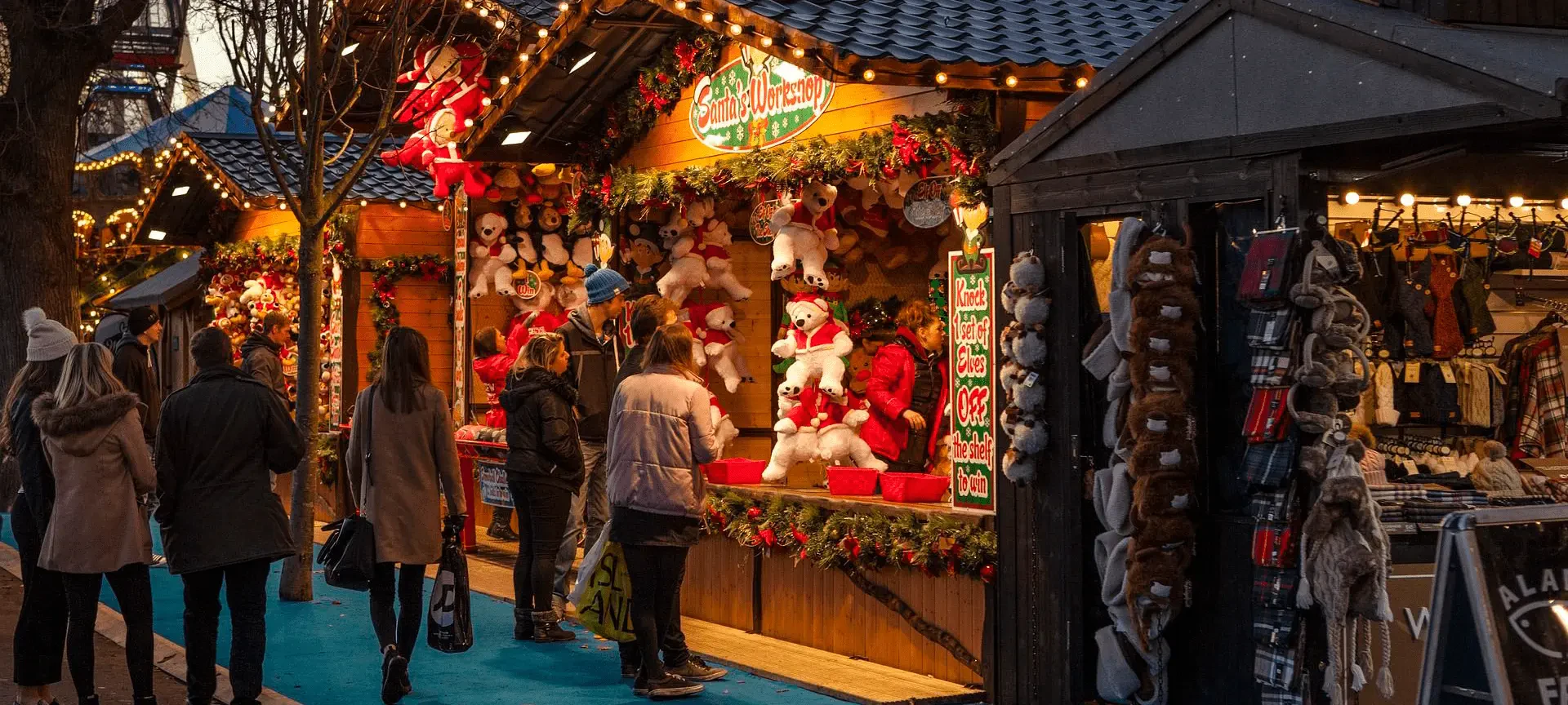 Puesto de mercado navideño decorado con ositos de peluche rojos y blancos y luces festivas, con compradores mirando cerca.