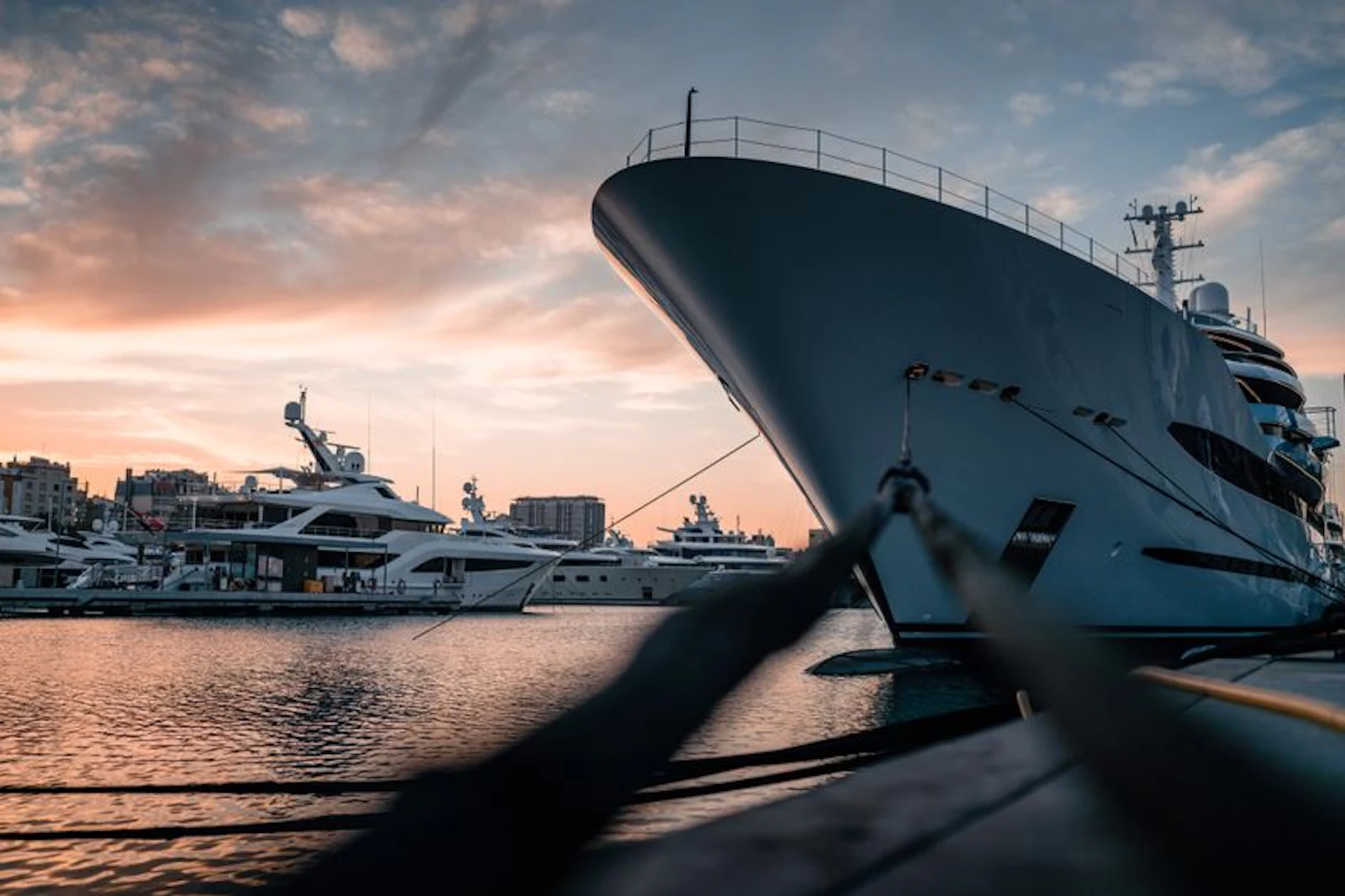 Large superyacht moored at marina at sunset with smaller yachts and waterfront buildings nearby.