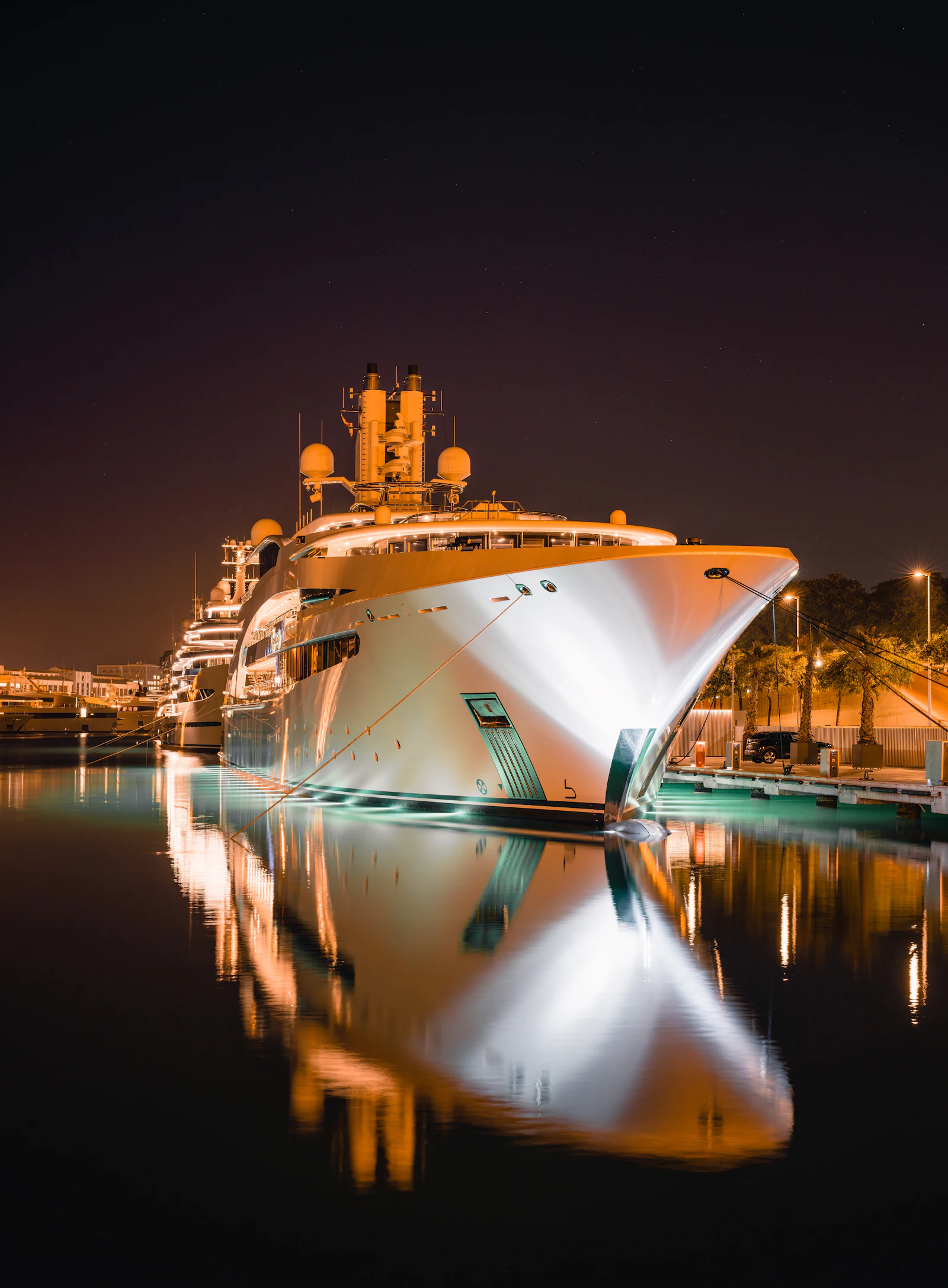 Illuminated superyacht moored at night in a marina with reflections in calm water.