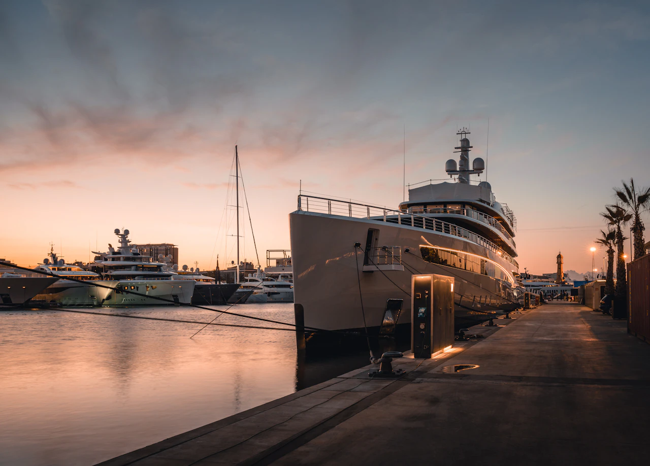 Large superyachts moored at a marina dock at sunset with palm trees and city buildings in the background.