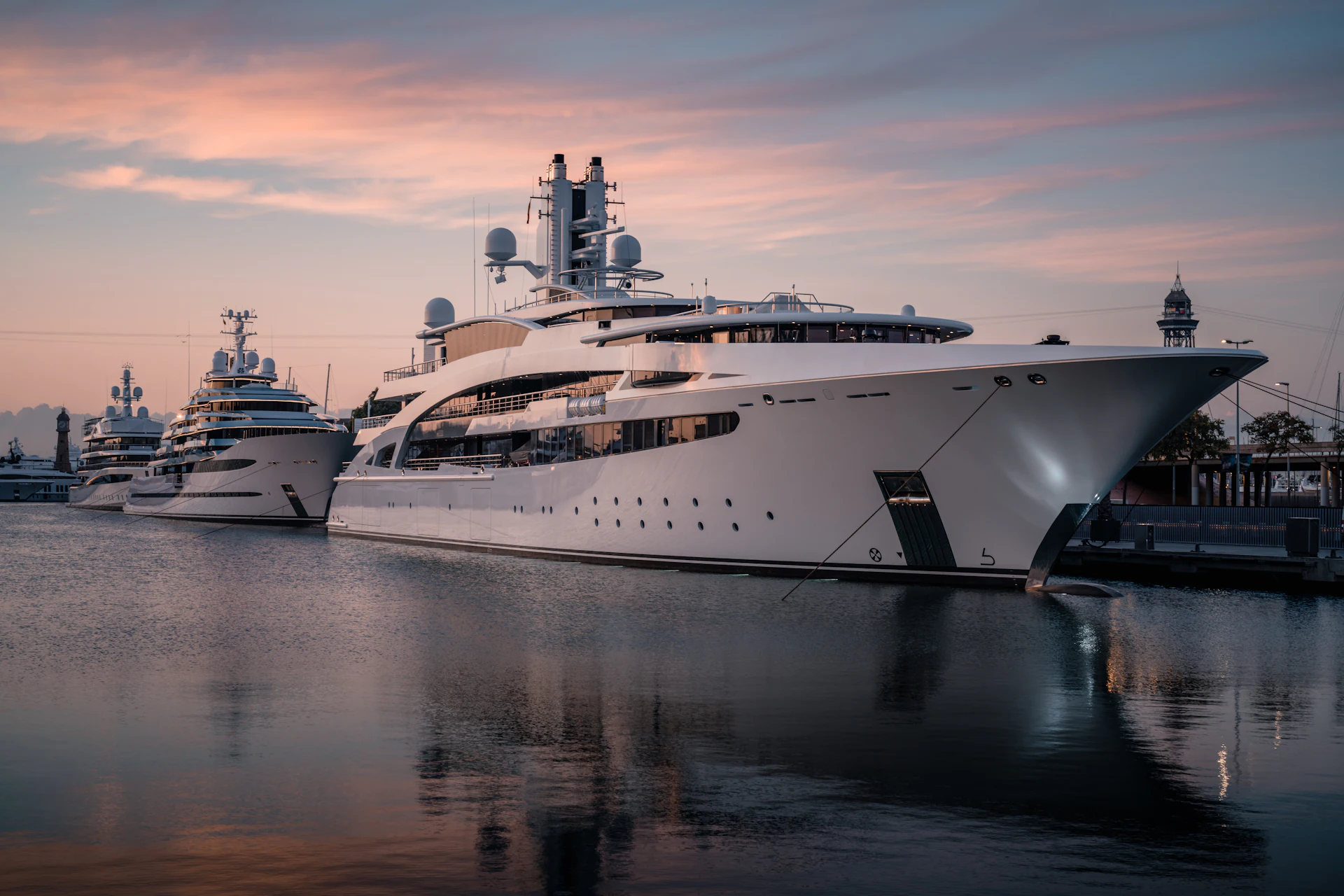Varios superyates amarrados en Marina Port Vell durante el atardecer con el perfil urbano de Barcelona de fondo.