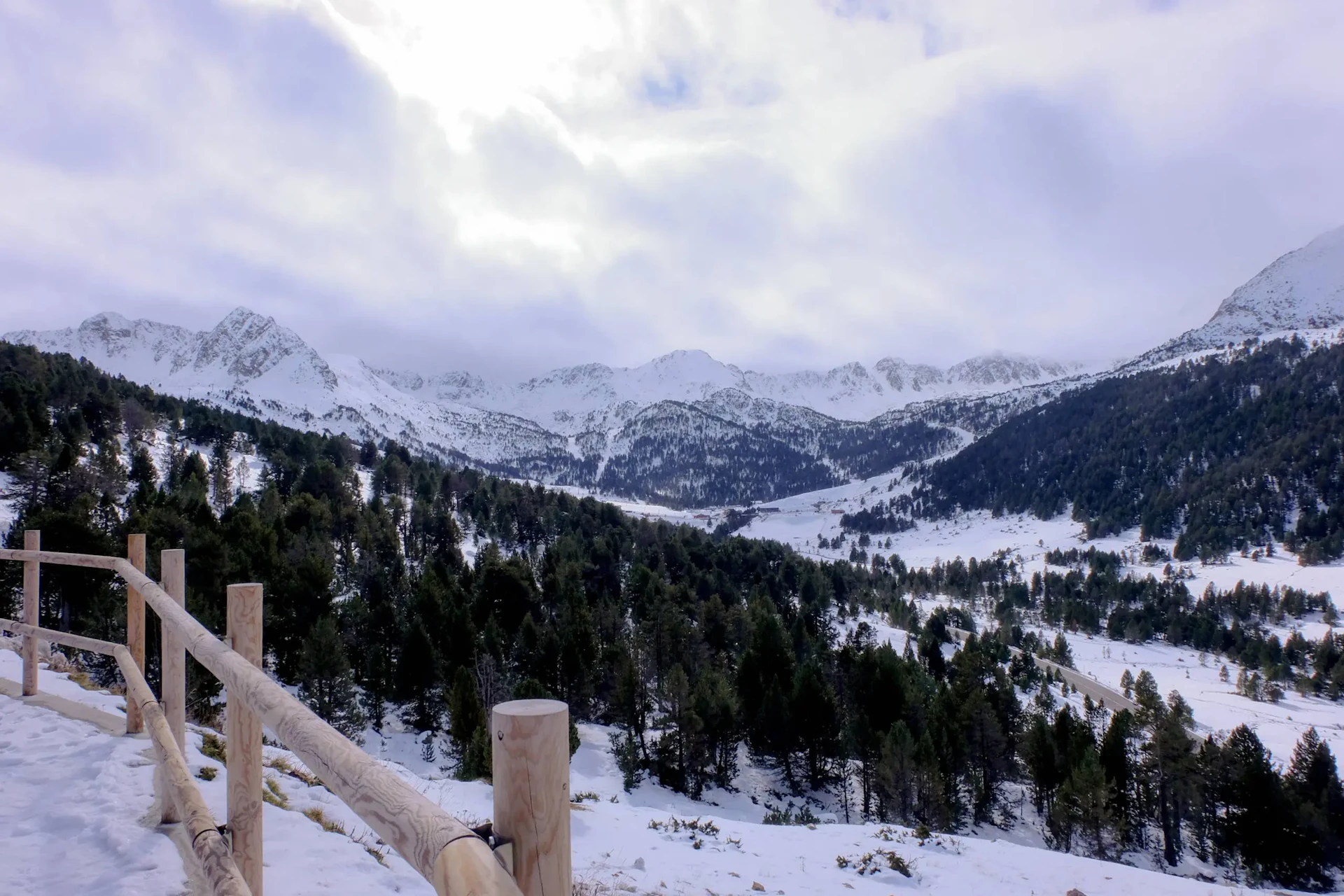 Valle de montaña cubierto de nieve con laderas boscosas y una valla de madera en primer plano.