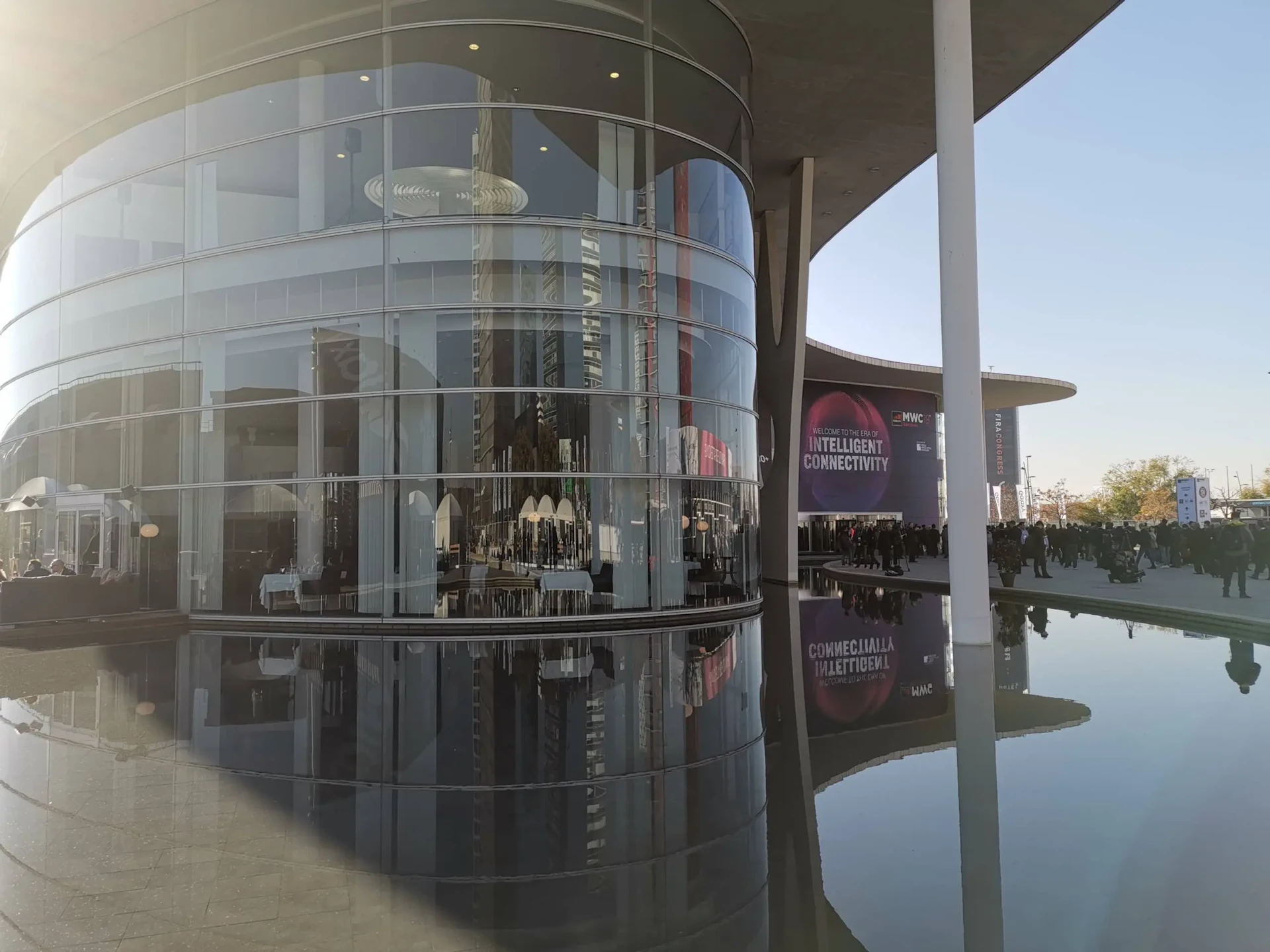 Modern curved glass building reflected in still water with people and banners visible in plaza.