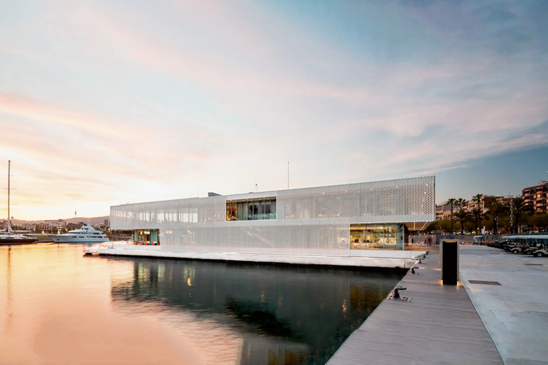 Edificio moderno blanco perforado en el muelle junto al agua con yates amarrados y el frente marítimo de Barcelona al fondo.