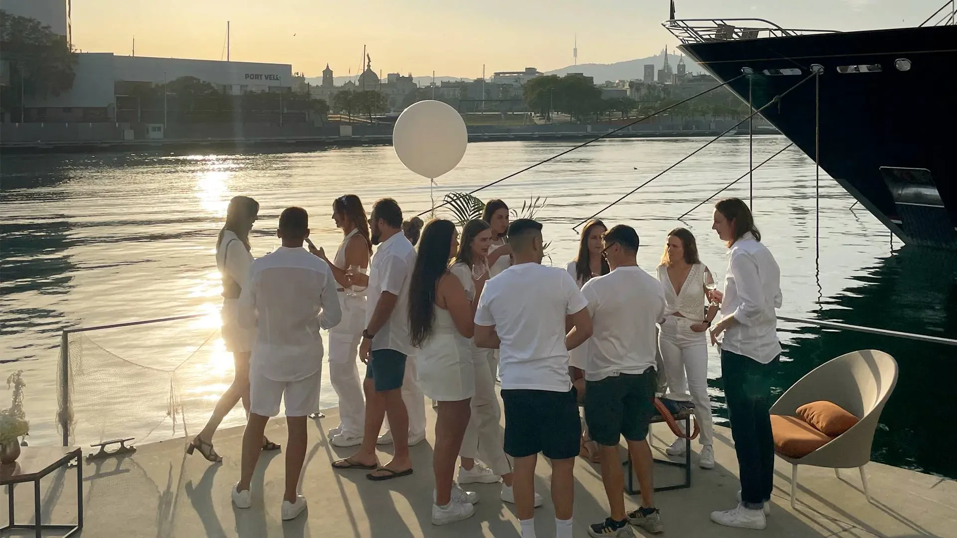 Group of people in white clothing gathered on a yacht deck at sunset with Barcelona cityscape in background.