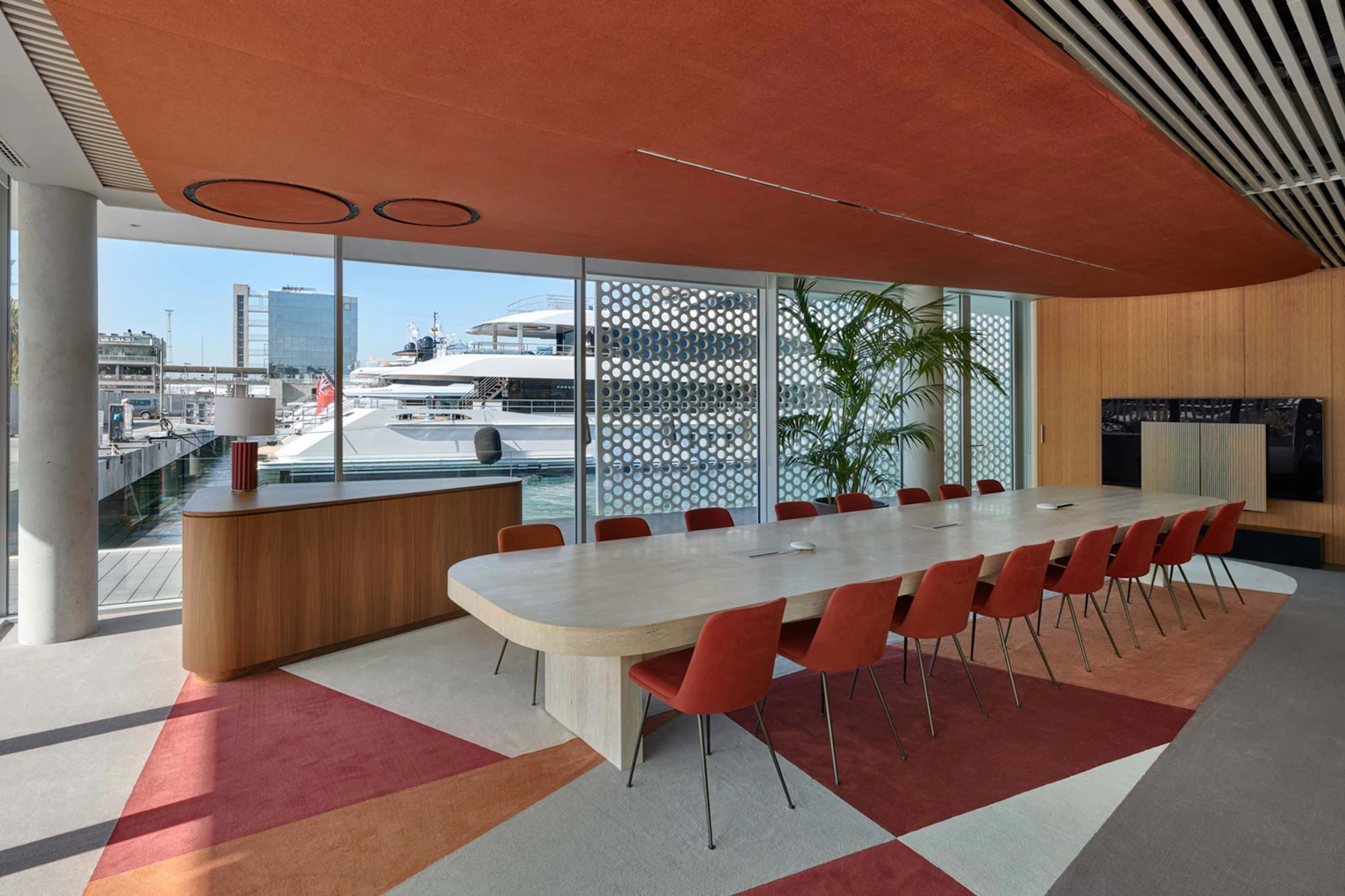 Sala de conferencias moderna con una larga mesa de piedra, sillas rojas y ventanales del suelo al techo con vistas a los superyates de Marina Port Vell.