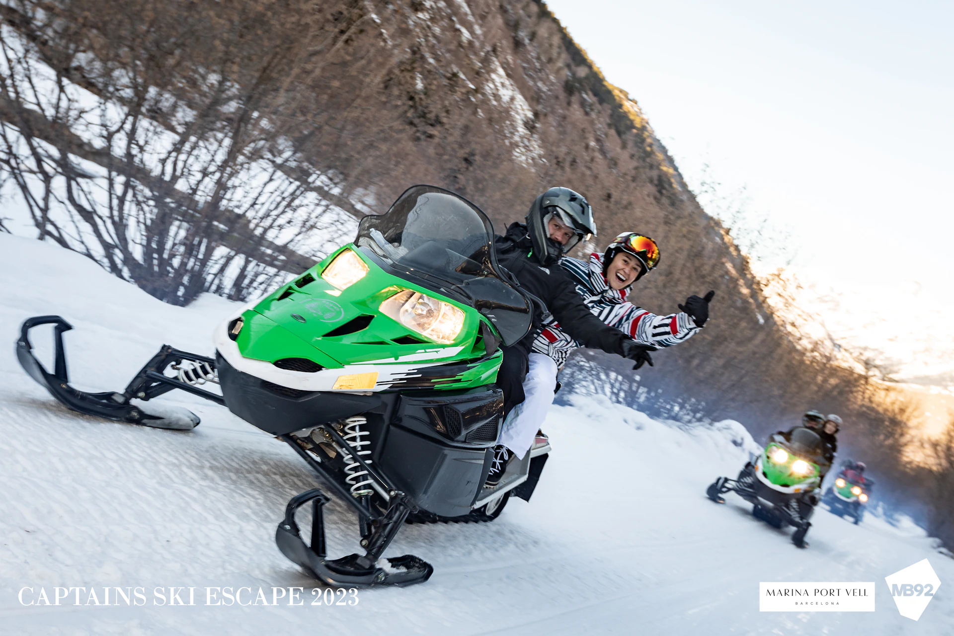 Dos personas montando una moto de nieve verde y negra sobre terreno nevado con un acantilado rocoso detrás.