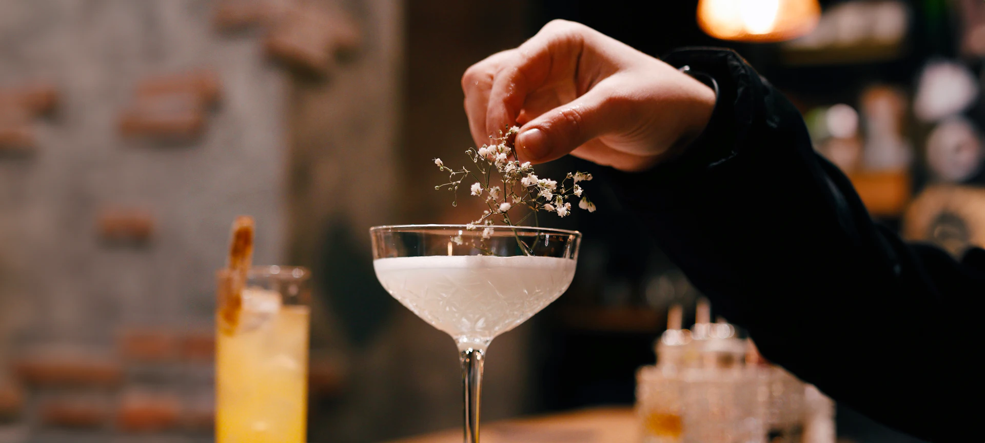 Bartender garnishing a cocktail with fresh herbs over a coupe glass at a bar.