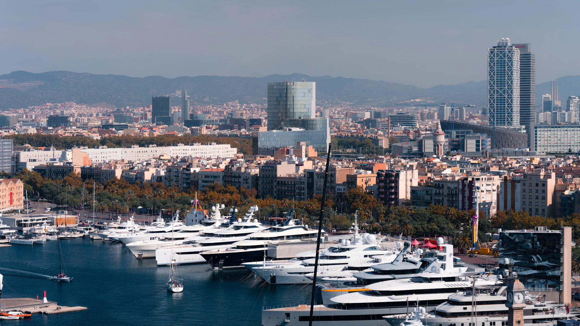 Varios superyates blancos amarrados en una marina de Barcelona con el perfil de la ciudad y montañas al fondo.