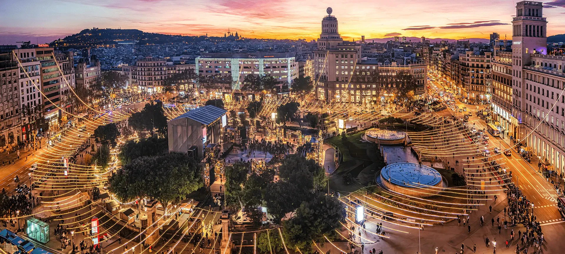 Barcelona cityscape at dusk with illuminated Plaza Reial, surrounding buildings, and Sagrada Familia in the distance.