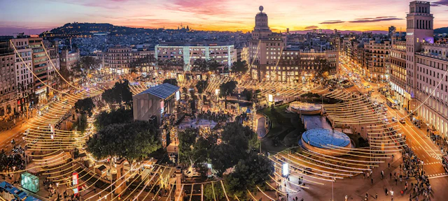 Paisaje urbano de Barcelona al anochecer con la Plaza Real iluminada, los edificios circundantes y la Sagrada Familia al fondo.