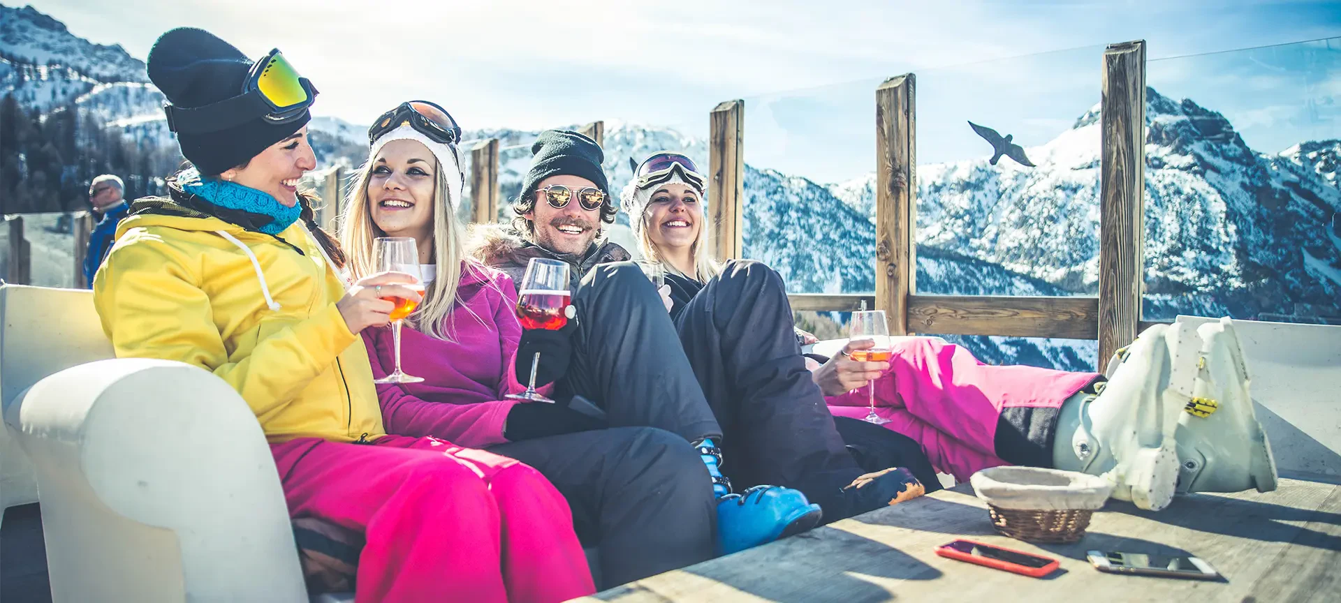 Cuatro personas con ropa de esquí de invierno disfrutan de unas bebidas en una terraza nevada de montaña con picos alpinos detrás de ellas.