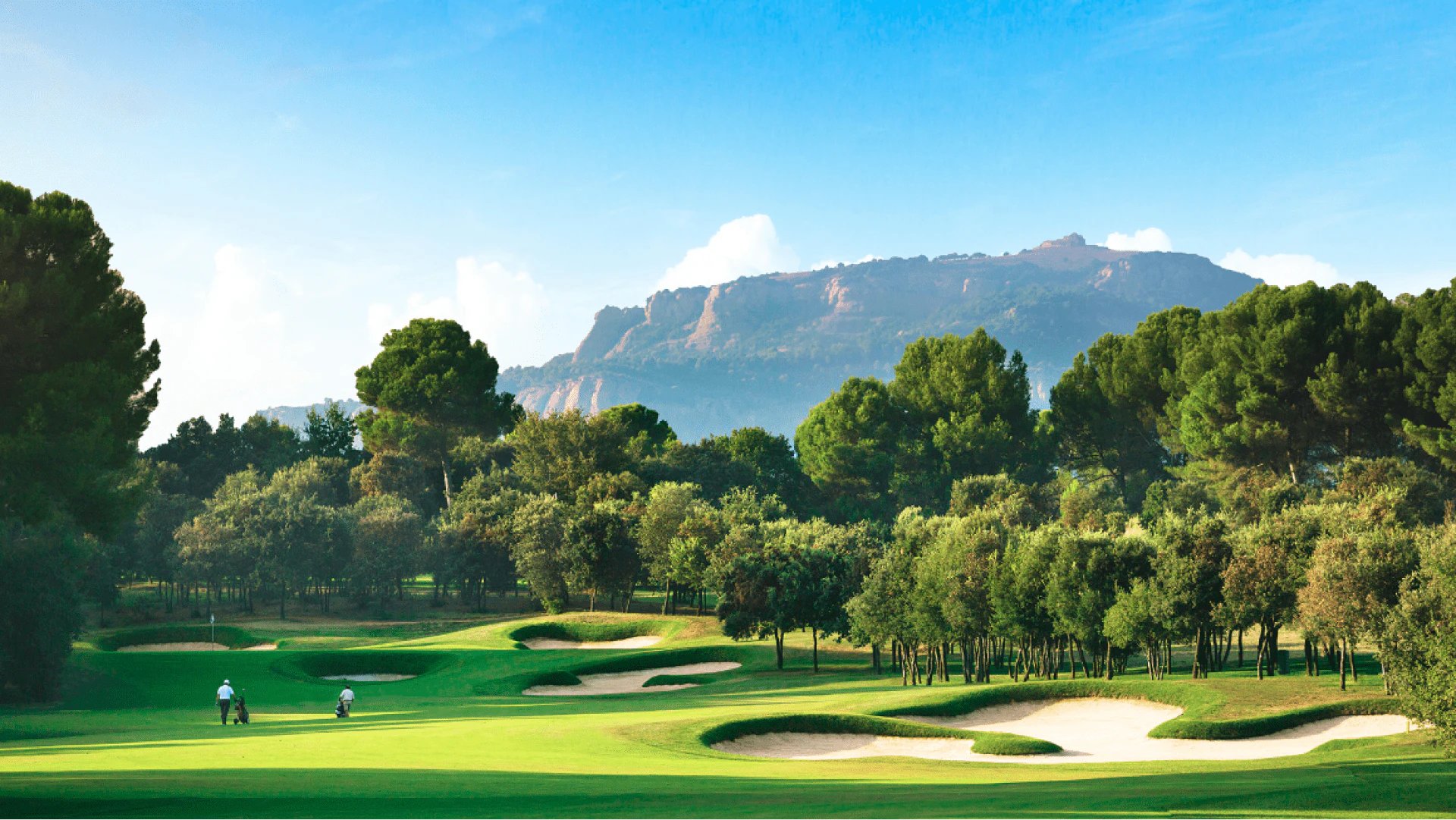 Golf course with sand bunkers and tree-lined fairways, mountains visible in the distance.