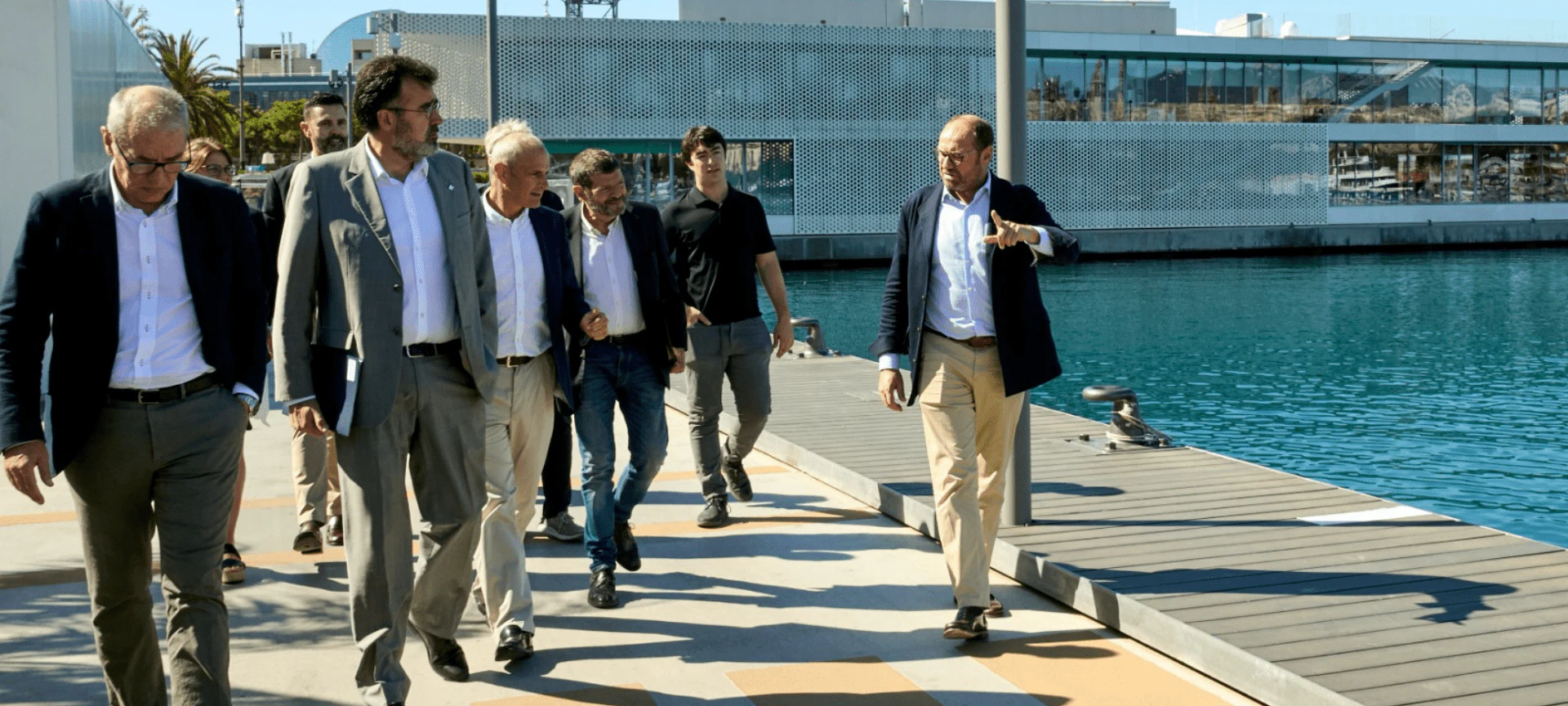 Grupo de hombres con traje caminando por un muelle de madera junto a aguas turquesas y modernos edificios frente al mar.