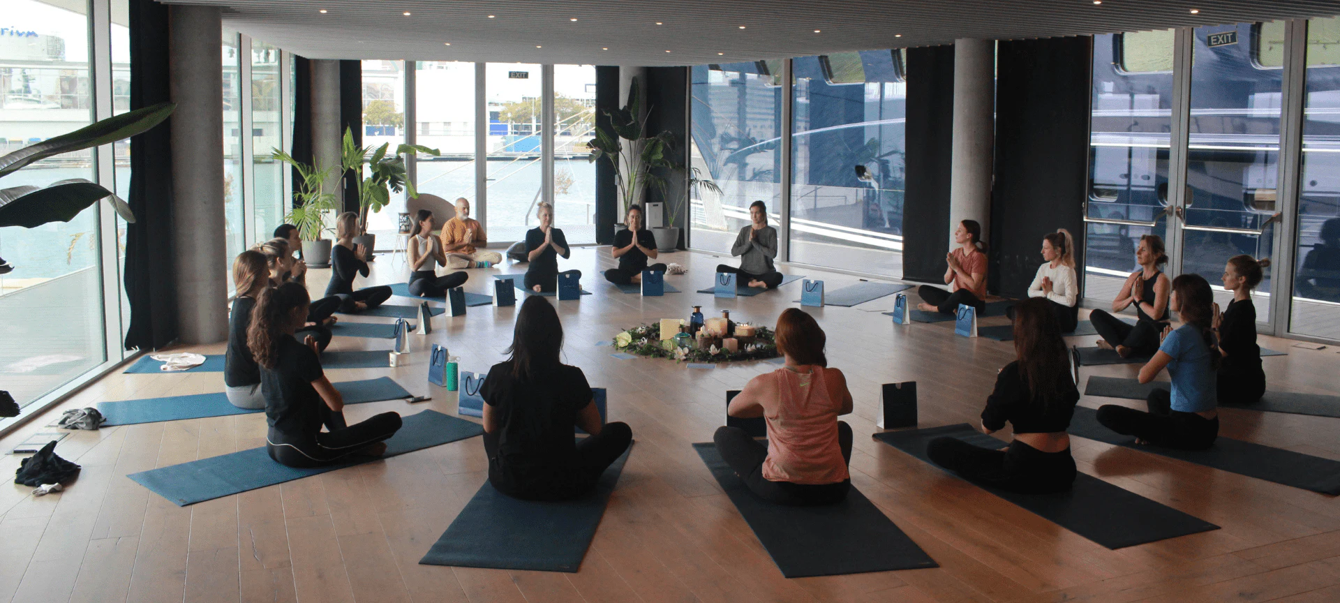 Grupo de personas sentadas en esterillas de yoga en un estudio junto al agua con vistas al puerto deportivo a través de grandes ventanales.