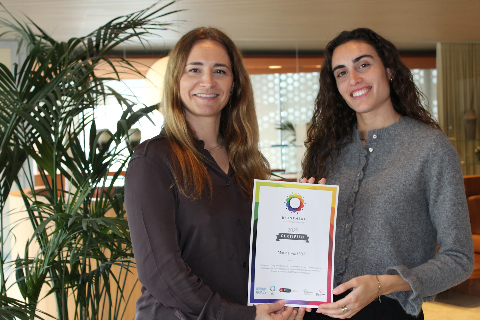 Two women holding a 2025 Biosphere certified award certificate in an indoor setting with plants.
