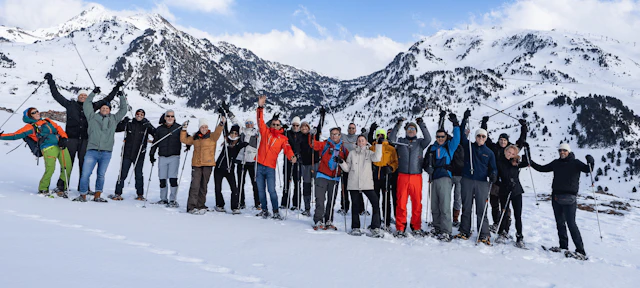 Large group of skiers and snowboarders posing together on a snowy mountain slope with snow-covered peaks in the background.