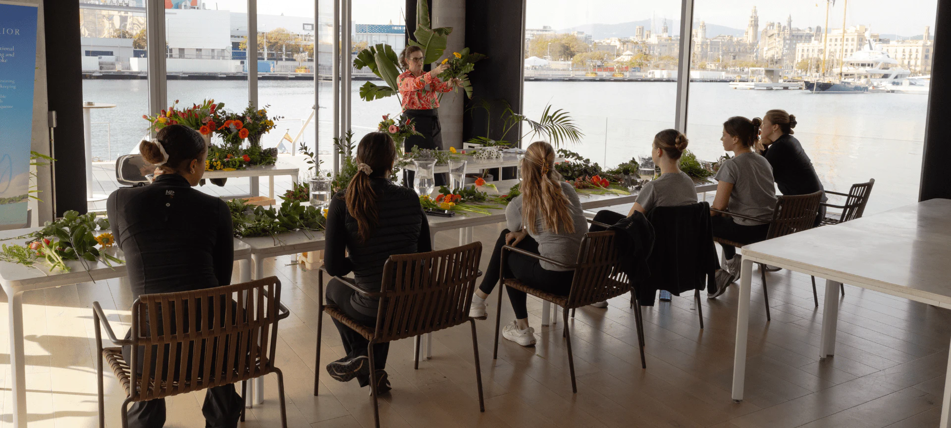 Grupo de personas sentadas alrededor de una mesa en un espacio luminoso frente al mar con vistas al paisaje urbano de Barcelona a través de grandes ventanales.