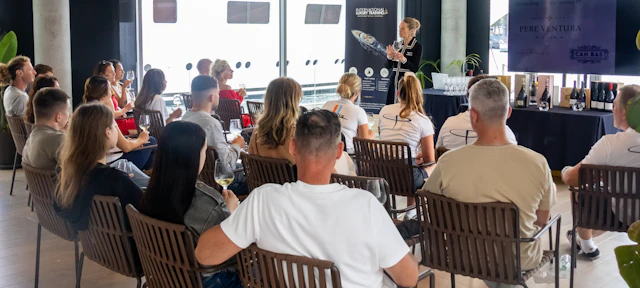 A woman presents to an audience seated in chairs overlooking a marina with superyachts docked behind.