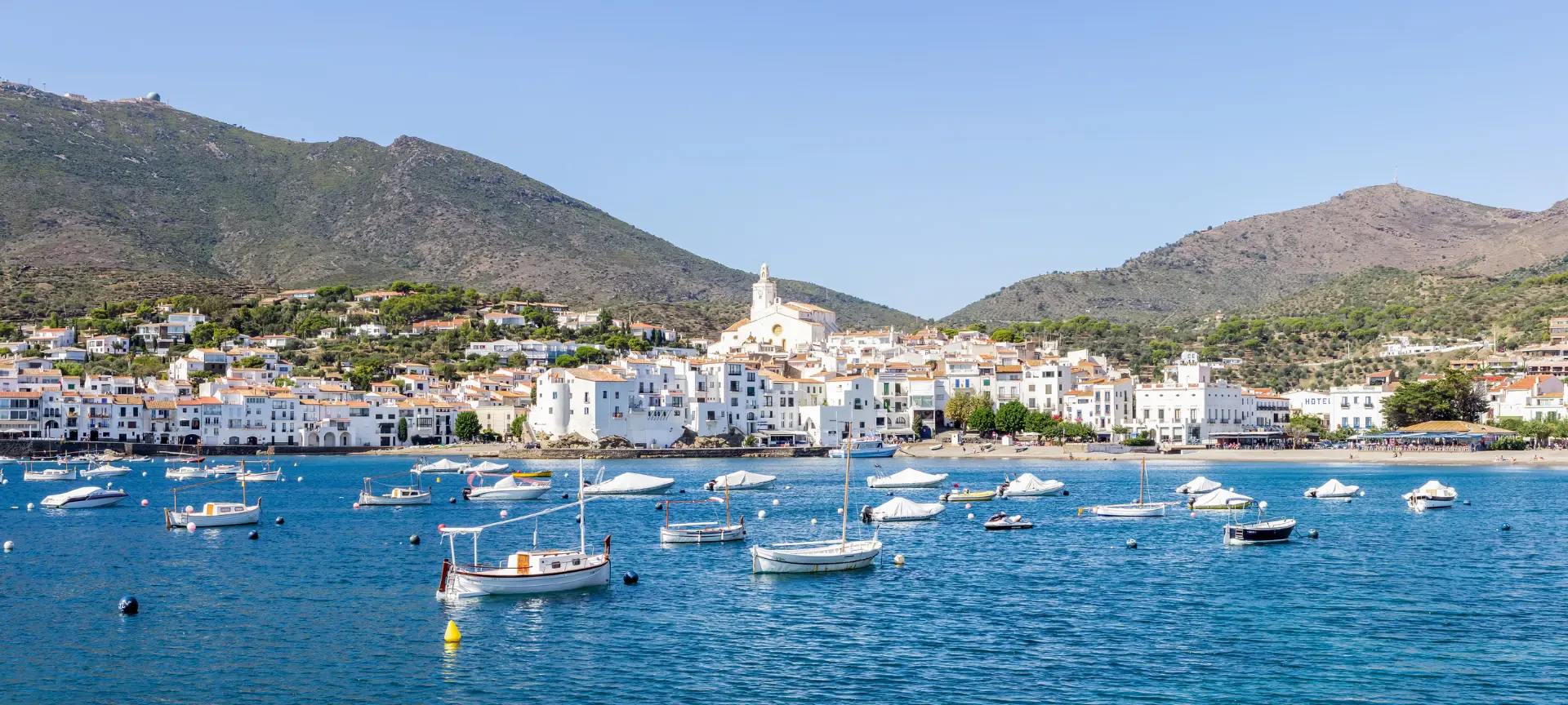 Pueblo costero mediterráneo con edificios blancos e iglesia entre colinas, barcos amarrados en una bahía turquesa.