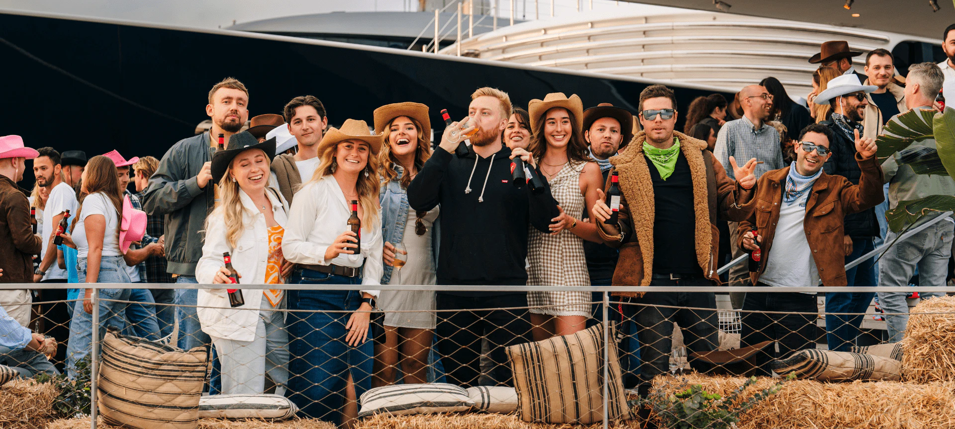 Invitados con sombreros de vaquero y atuendo western celebran en un muelle de la marina junto a un gran yate.