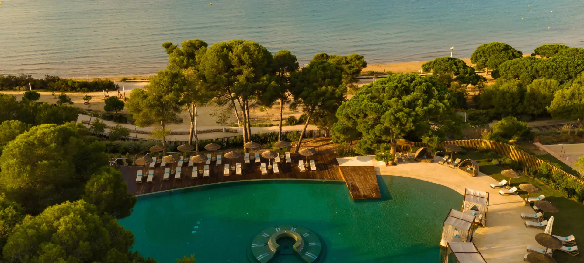 Vista aérea de una piscina frente a la playa con tumbonas y sombrillas, rodeada de pinos y costa mediterránea.