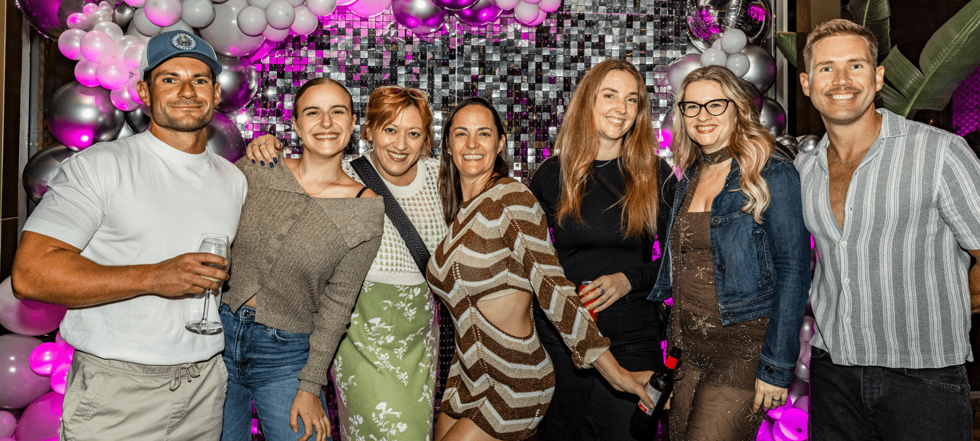 Siete personas posando juntas en un evento de interior con decoración de globos rosas y plateados detrás de ellas.