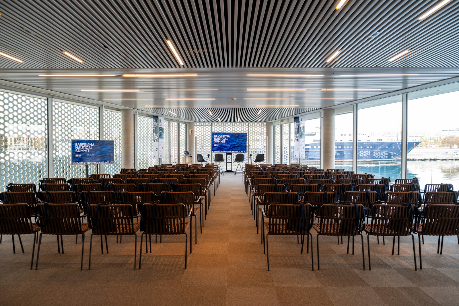 Sala de conferencias moderna con filas de sillas negras orientadas hacia un escenario y una pantalla azul con vistas a un puerto deportivo.